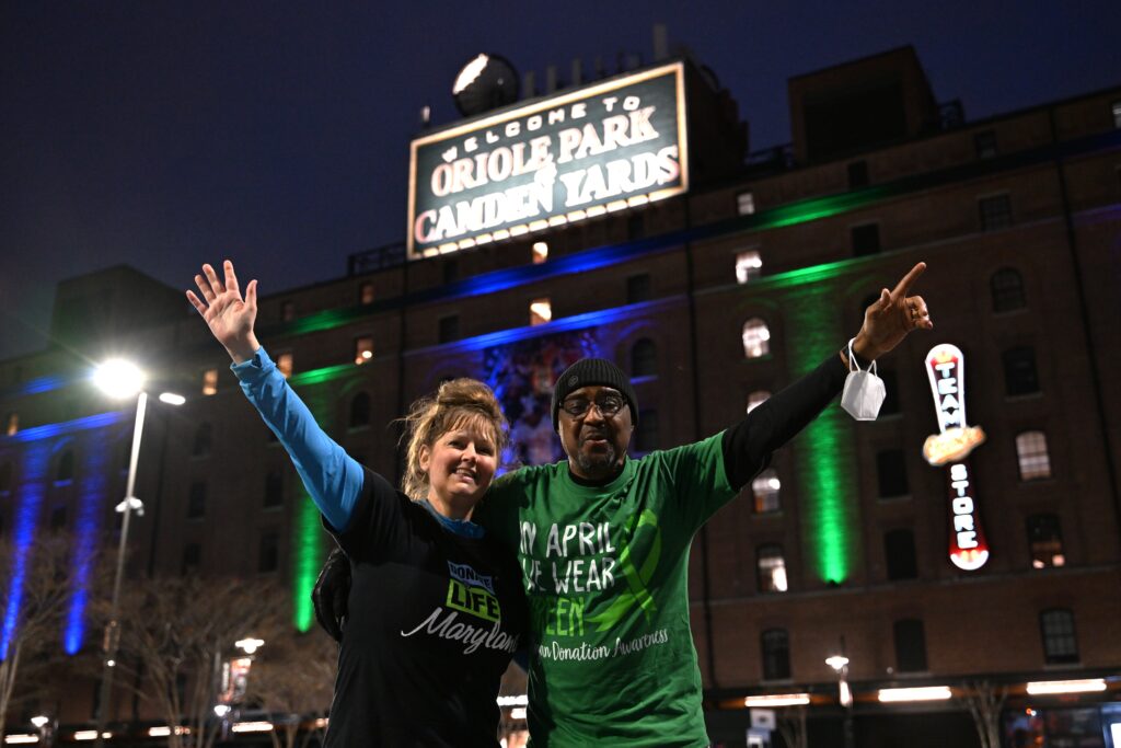 A man and a woman stand in front of the blue and green lights lit at Camden Yards in honor of April being National Donate Life Month.