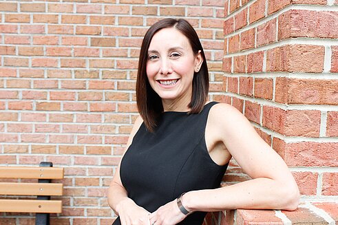 Vice President and Chief Human Resources Officer, Jaclyn Quackenbush leans against a brick wall outside of Infinite Legacy's office smiling.
